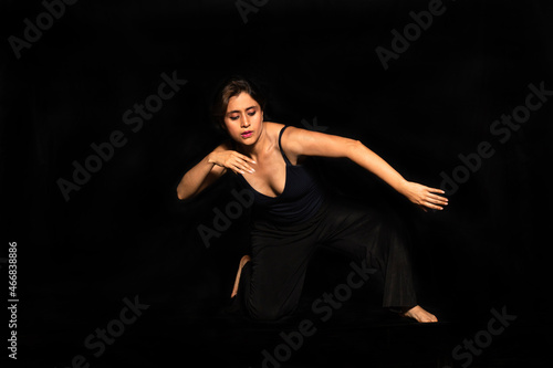 Full body portrait of a latin woman posing with one knee on the ground and moving her arms isolated on black background. Female body expression concept. © Julian