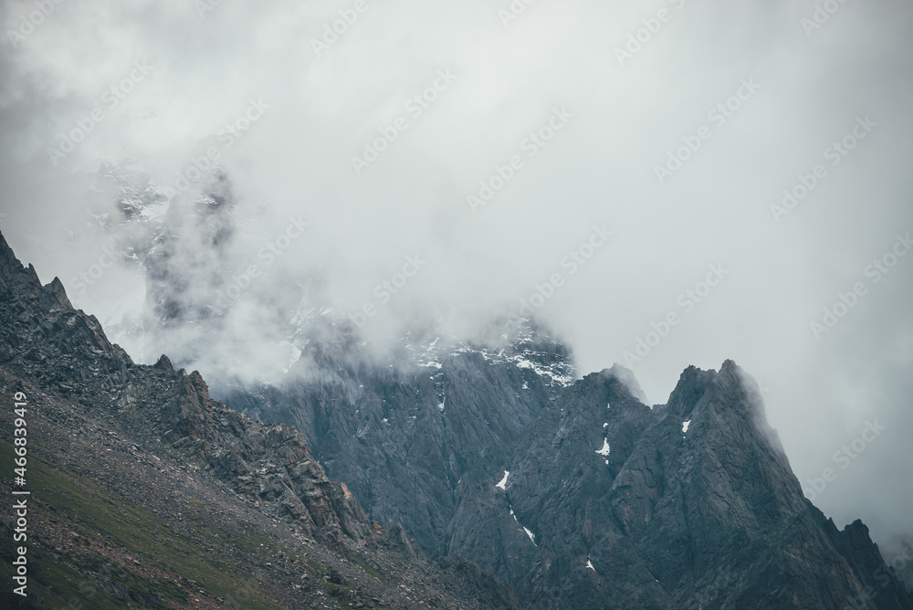 Dark atmospheric surreal landscape with dark rocky mountain top in low clouds in gray cloudy sky. Gray low cloud on high pinnacle. High black rock with snow in low clouds. Surrealist gloomy mountains. Dark atmospheric surreal landscape with dark rocky mountain top in low clouds in gray cloudy sky. Gray low cloud on high pinnacle. High black rock with snow in low clouds. Surrealist gloomy mountains.