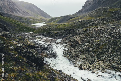 Atmospheric landscape with mountain lake and mountain creek among moraines in rainy weather. Bleak overcast scenery with milky river and lake among rocks. Gloomy view to milk mountain river and lake.