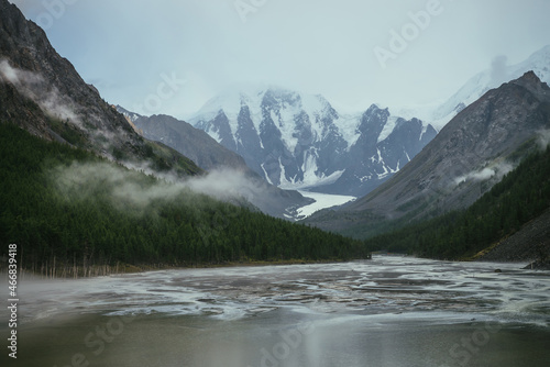 Atmospheric alpine landscape with mountain lake with streams from snowy mountains in overcast weather. Gloomy mountain scenery with green lake with rainy circles and low clouds in mountain valley. © Daniil