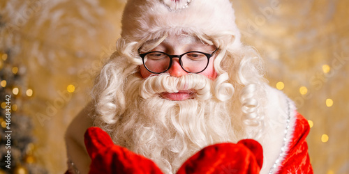Portrait of santa clois with glasses blowing on mittens against the background of garlands. Selective focus. Beautiful christmas card photo