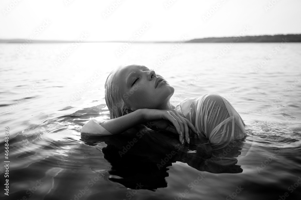 Fototapeta premium Beautiful blonde woman short hair lies in water on sea shore, pond. Art portrait of a relaxing woman in the water on shore