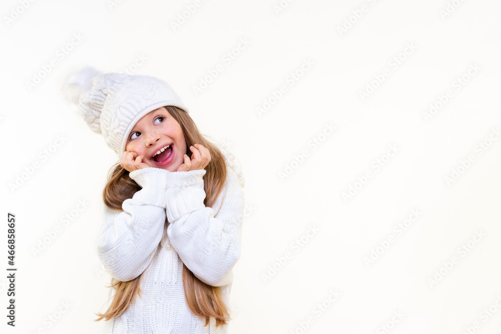 A little girl in a warm sweater . The girl looks up in surprise and holds on to her hat.