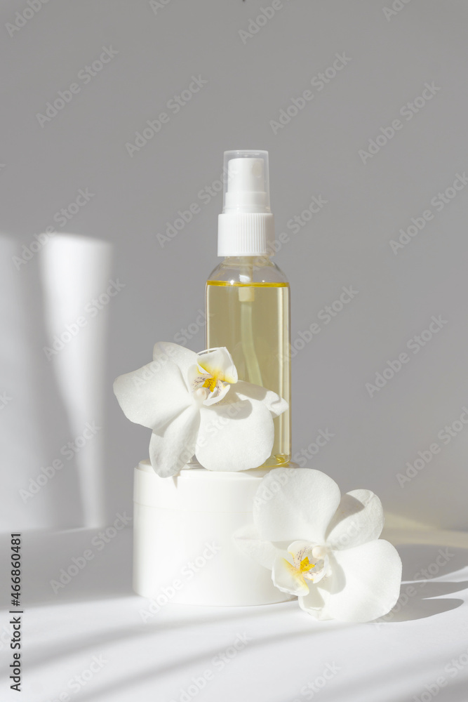Mock-up of glass bottle with essential oil, standing on white jar with cream, surrounded by orchid flowers. Vertical white background with morning sun rays. Concept of beauty industry, spa treatments Mock-up of glass bottle with essential oil, standing on white jar with cream, surrounded by orchid flowers. Vertical white background with morning sun rays. Concept of beauty industry, spa treatments