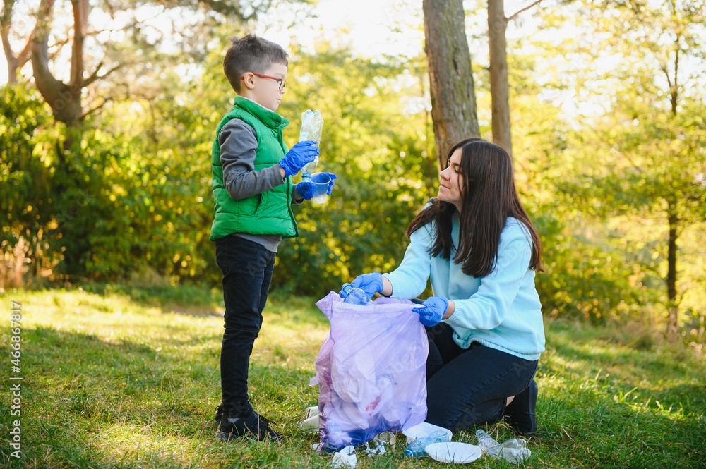 Fototapeta premium Smiling boy picking up trash in the park with his mother. Volunteer concept.