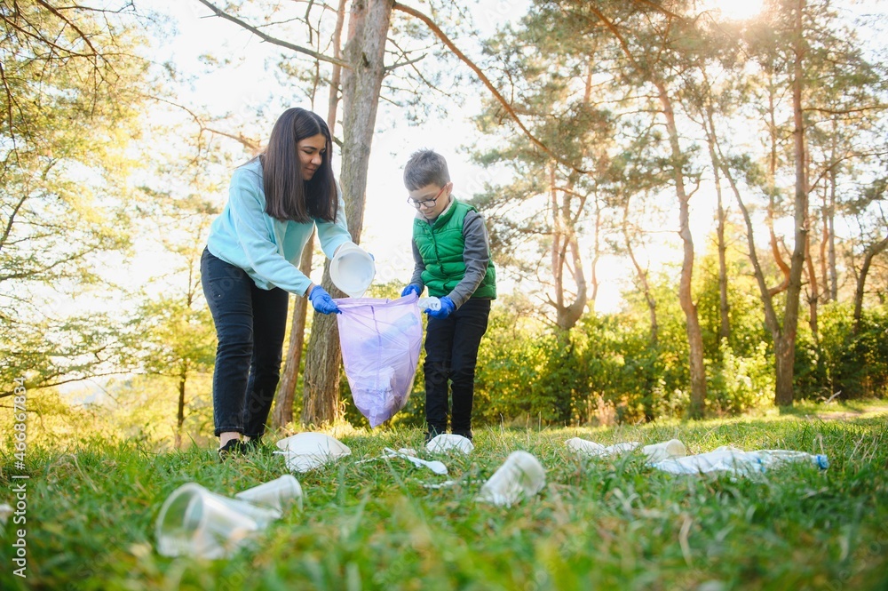 Fototapeta premium Smiling boy picking up trash in the park with his mother. Volunteer concept.