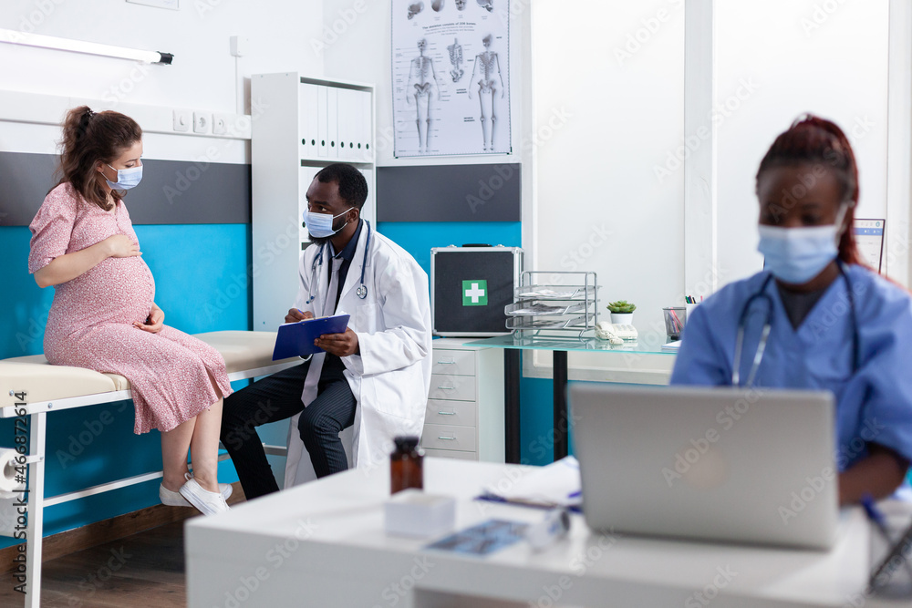 African american medic talking to pregnant woman in cabinet while wearing face masks. Specialist doing consultation with patient expecting child for healthcare during coronavirus pandemic.