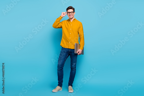 Full length photo of clever brunet ceo man hold laptop wear yellow shirt eyewear jeans sneakers isolated on blue color background