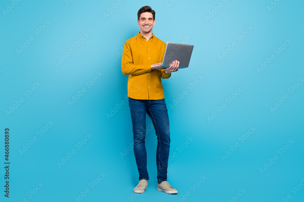 Photo of cheerful freelancer guy hold netbook typing wear yellow shirt jeans shoes isolated blue color background Photo of cheerful freelancer guy hold netbook typing wear yellow shirt jeans shoes isolated blue color background