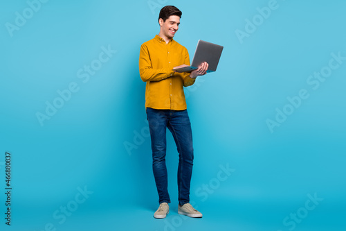 Full size photo of smart brunet man look laptop wear yellow shirt jeans footwear isolated on blue color background
