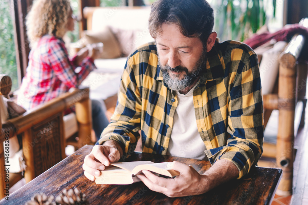 Adult man reading a book at home sitting on the wooden table with wife woman in background. Happy caucasian couple of people enjoying leisure time indoor together with relax Adult man reading a book at home sitting on the wooden table with wife woman in background. Happy caucasian couple of people enjoying leisure time indoor together with relax