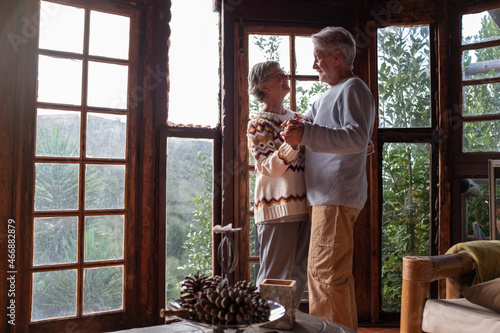 Happy senior couple in love dance at home agains a big windows view with nature woods outside. Romance leisure activity mature man and woman elderly retired lifestyle photo