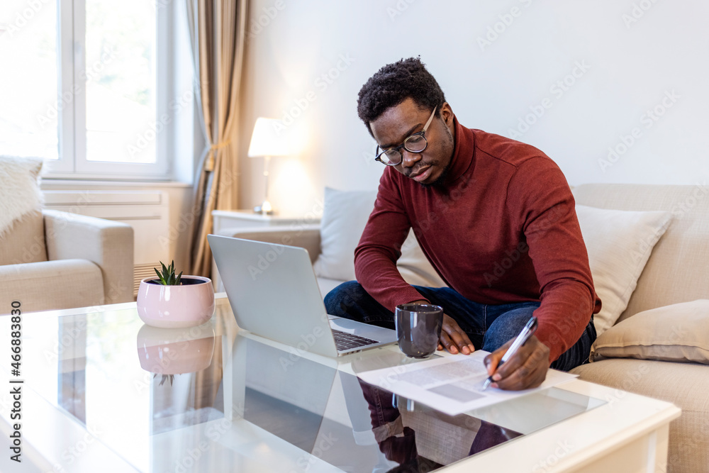 African-American male student or worker in glasses is watching online lectures or webinars and writing notes in a notebook. Concentrated dark skinned copywriter in eyewear typing text of publication 