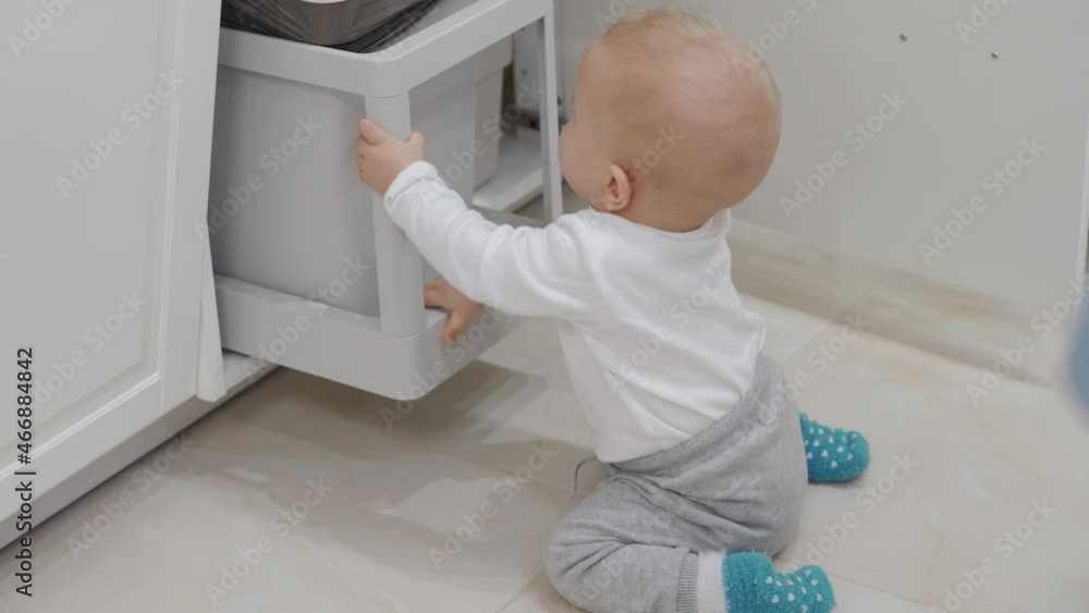 Home has many potential hazards for a child, baby opening cabinet grabbing and playing with pull out trash can under kitchen counter, safety at home for kids. High quality 4k footage Home has many potential hazards for a child, baby opening cabinet grabbing and playing with pull out trash can under kitchen counter, safety at home for kids. High quality 4k footage