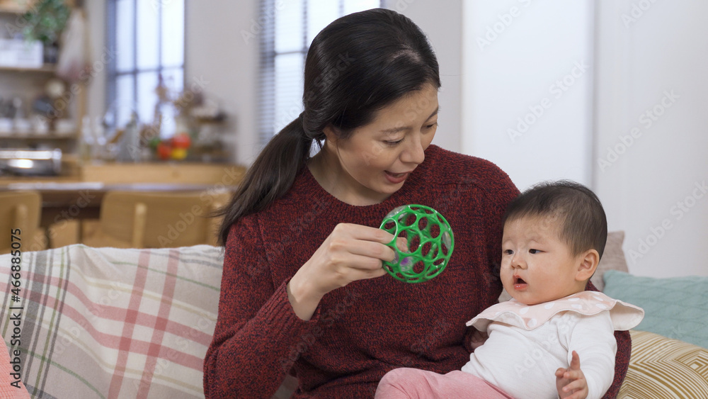 happy asian mother is playing with her lovely baby daughter in arms by shaking a toy ball to her and putting it her hands on sofa at home. happy asian mother is playing with her lovely baby daughter in arms by shaking a toy ball to her and putting it her hands on sofa at home.