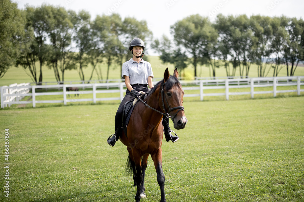 Female horseman riding brown Thoroughbred horse on green meadow in countryside. Concept of rural resting and leisure. Green tourism. Young smiling european woman. Beautiful landscape at sunny day Female horseman riding brown Thoroughbred horse on green meadow in countryside. Concept of rural resting and leisure. Green tourism. Young smiling european woman. Beautiful landscape at sunny day