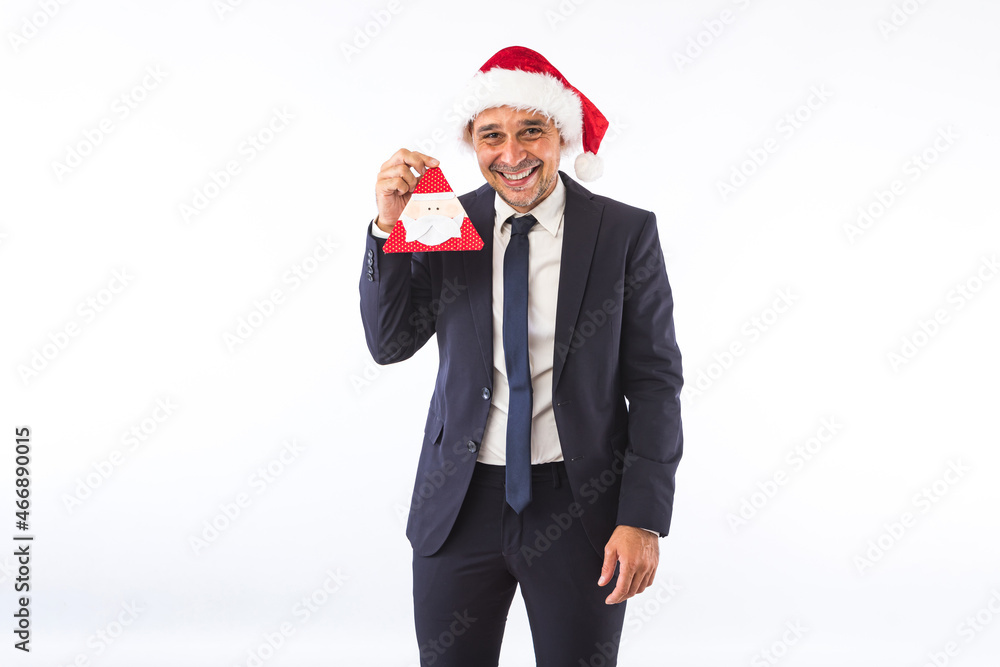 Businessman dressed in suit, tie and Christmas hat of Santa Claus, smiling, holding a Santa ornament, on white background. Christmas celebration concept. Businessman dressed in suit, tie and Christmas hat of Santa Claus, smiling, holding a Santa ornament, on white background. Christmas celebration concept.