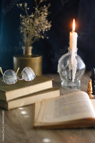Old books  reading glasses  vintage chess pieces  lit candle and vase with gypsophila flowers. Dark academia concept. Selective focus.
