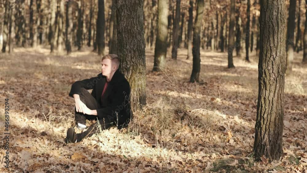 Portrait of handsome man in black coat sitting near the tree in autumn forest on sunny day Portrait of handsome man in black coat sitting near the tree in autumn forest on sunny day