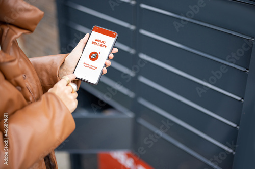 Cropped image of woman putting parcel to cell of automatic post terminal and showing smartphone with smart delivery app. Concept of modern shipping and logistics