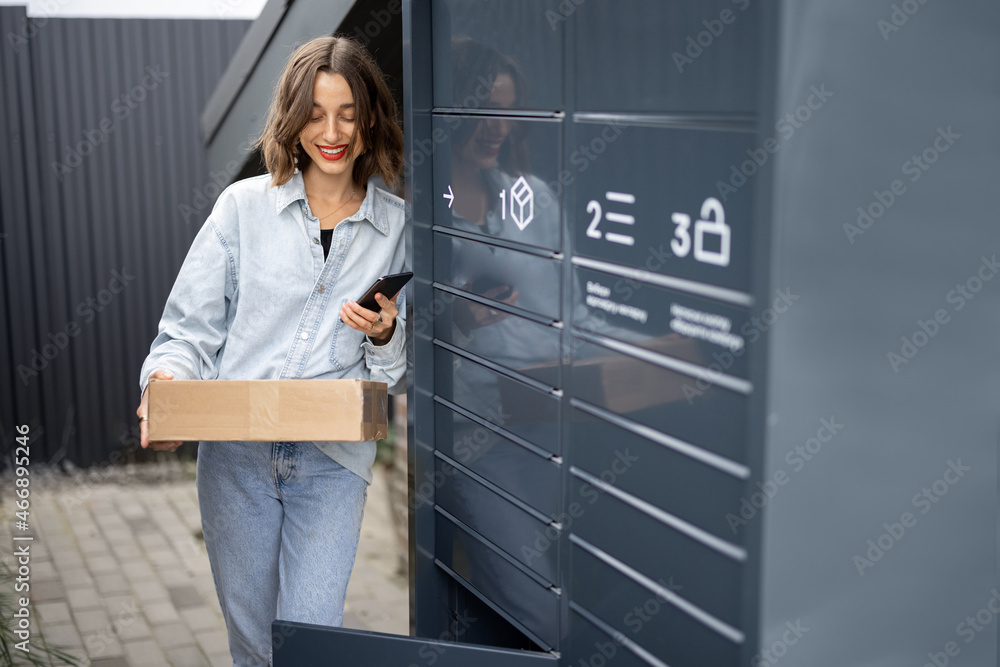 Young caucasian woman with parcel near automatic post terminal. Smiling girl holding smartphone and looking away on city street. Concept of smart delivery. Idea of modern shipping and logistics