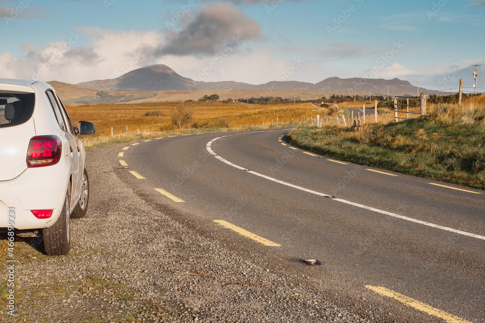 White car parked off small country road. Beautiful nature scenery with fields and Croagh Patrick mountain. County Mayo, Ireland. Travel and road trip concept. Blue cloudy sky.