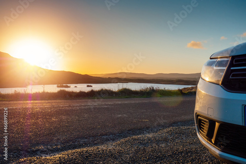 Car parked by a small lake with blue water and wild fields. Beautiful sun rise scene in Connemara, county Galway, Ireland. Warm and cool tones. Haze over mountains in the background.