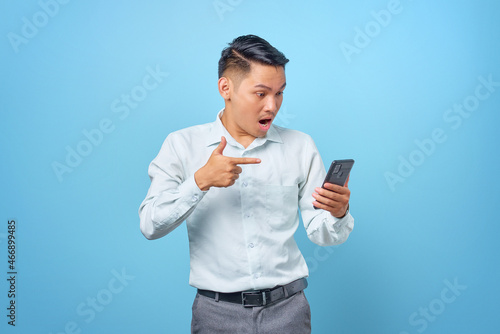 Shocked young handsome businessman pointing finger at smartphone on blue background