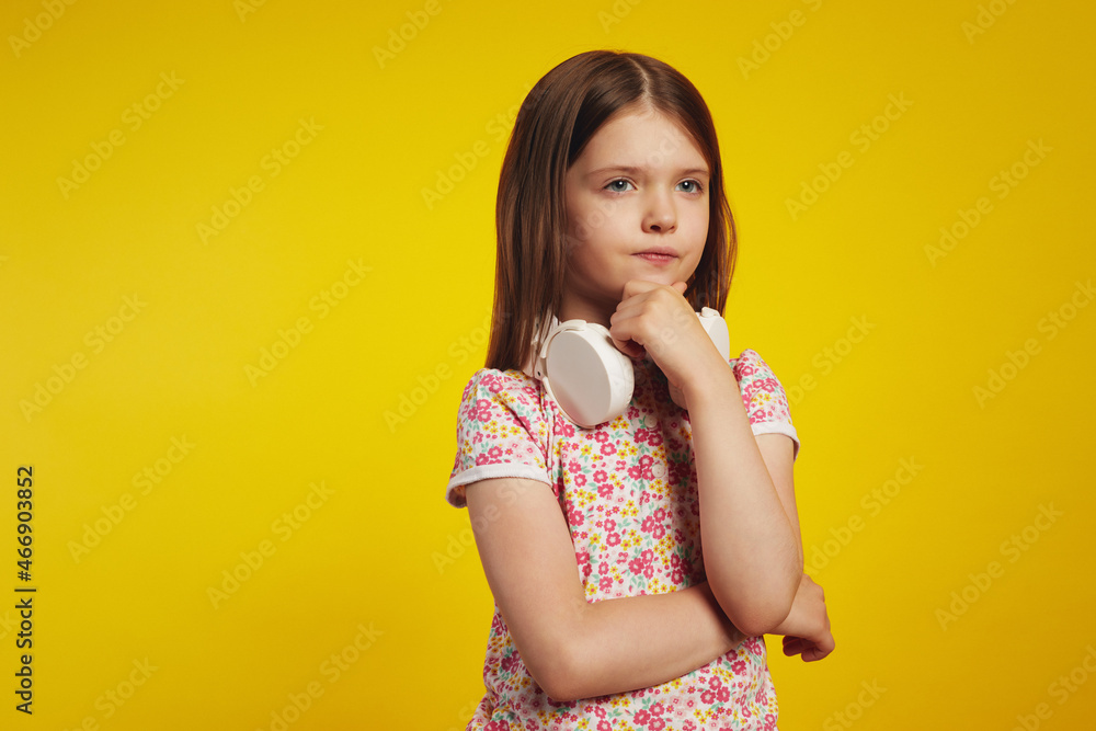 Thoughtful little kid in summer clothes and white headphones on neck touching chin while looking away, isolated against yellow background Thoughtful little kid in summer clothes and white headphones on neck touching chin while looking away, isolated against yellow background