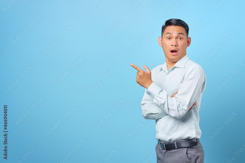 Shocked young handsome businessman pointing finger away at copy space on blue background Shocked young handsome businessman pointing finger away at copy space on blue background