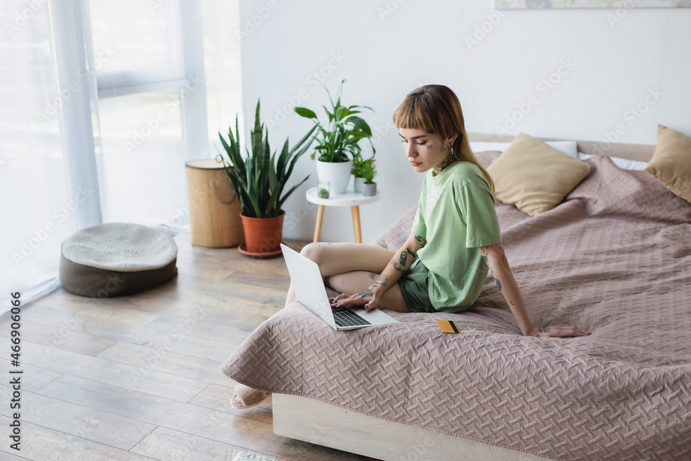 woman with tattooed body typing on laptop while sitting on bed near credit card woman with tattooed body typing on laptop while sitting on bed near credit card