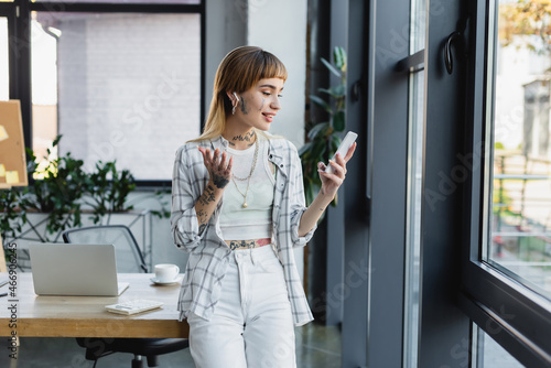 smiling businesswoman with tattoo gesturing during video call on smartphone in office