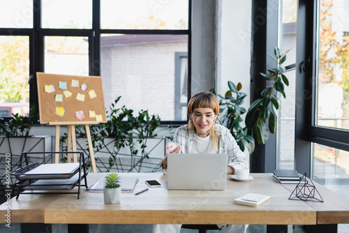 positive tattooed woman gesturing during video chat on laptop at workplace