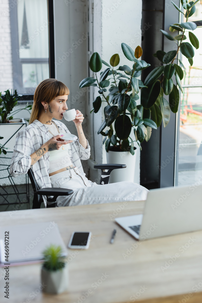 tattooed businesswoman drinking coffee while sitting near work desk and potted plant tattooed businesswoman drinking coffee while sitting near work desk and potted plant