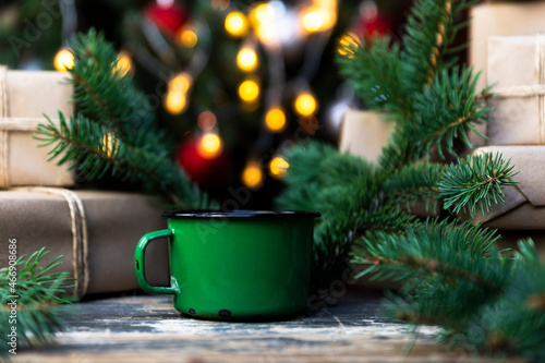 Coffee in a tin mug on the rustic table with wrapped gifts, Christmas tree lights and spruce branches in the background