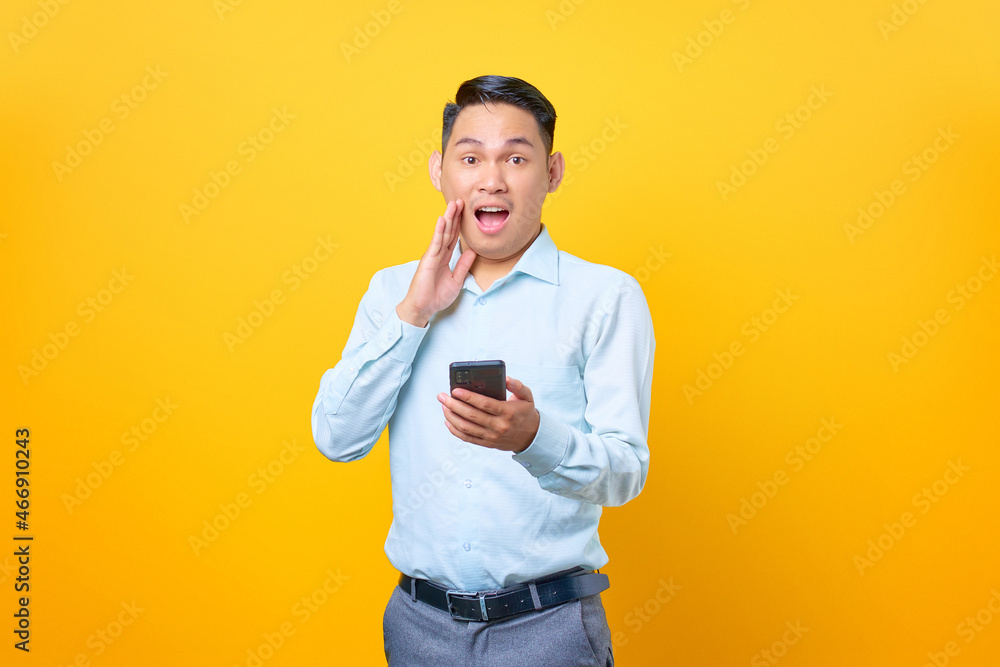 Shocked young handsome businessman holding smartphone and looking camera on yellow background Shocked young handsome businessman holding smartphone and looking camera on yellow background
