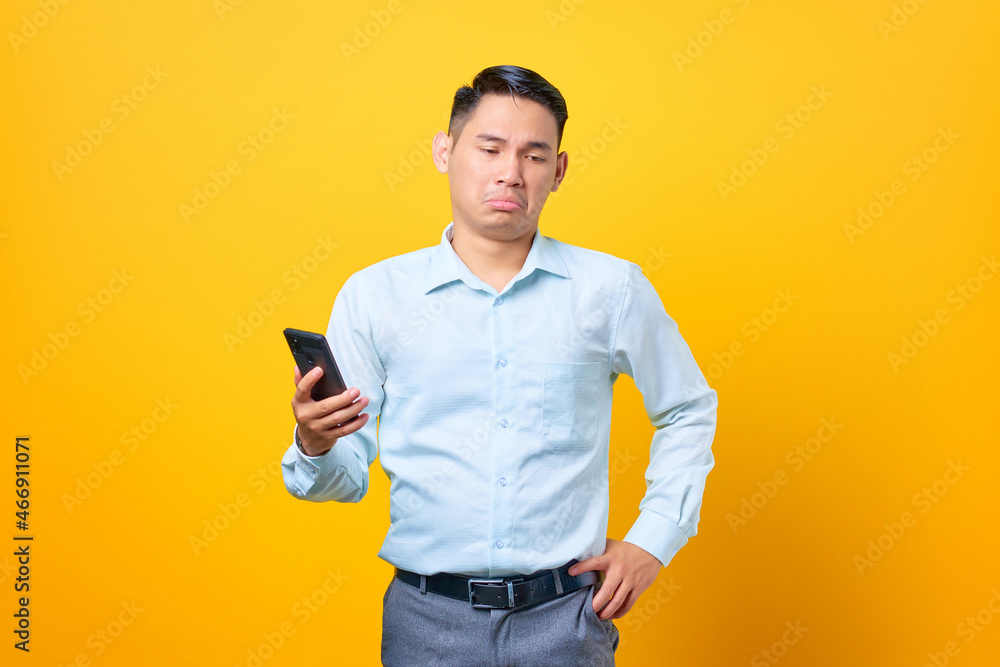 Sad young handsome businessman holding smartphone on yellow background Sad young handsome businessman holding smartphone on yellow background