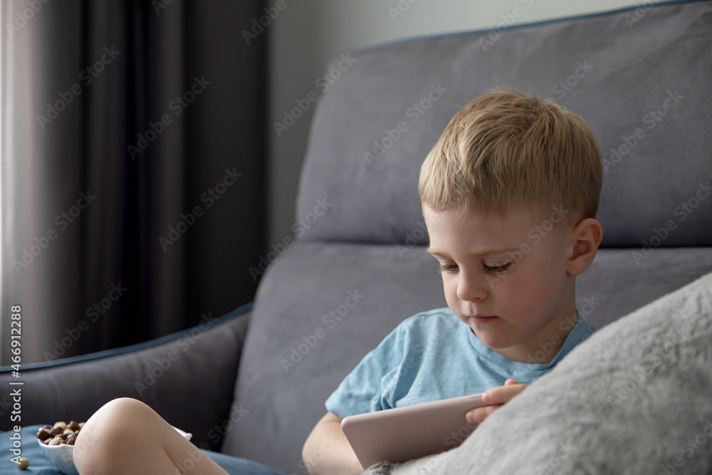 Cute little boy blonde looks at the smartphone sitting on the couch at home. Concept online education, learning, games Cute little boy blonde looks at the smartphone sitting on the couch at home. Concept online education, learning, games