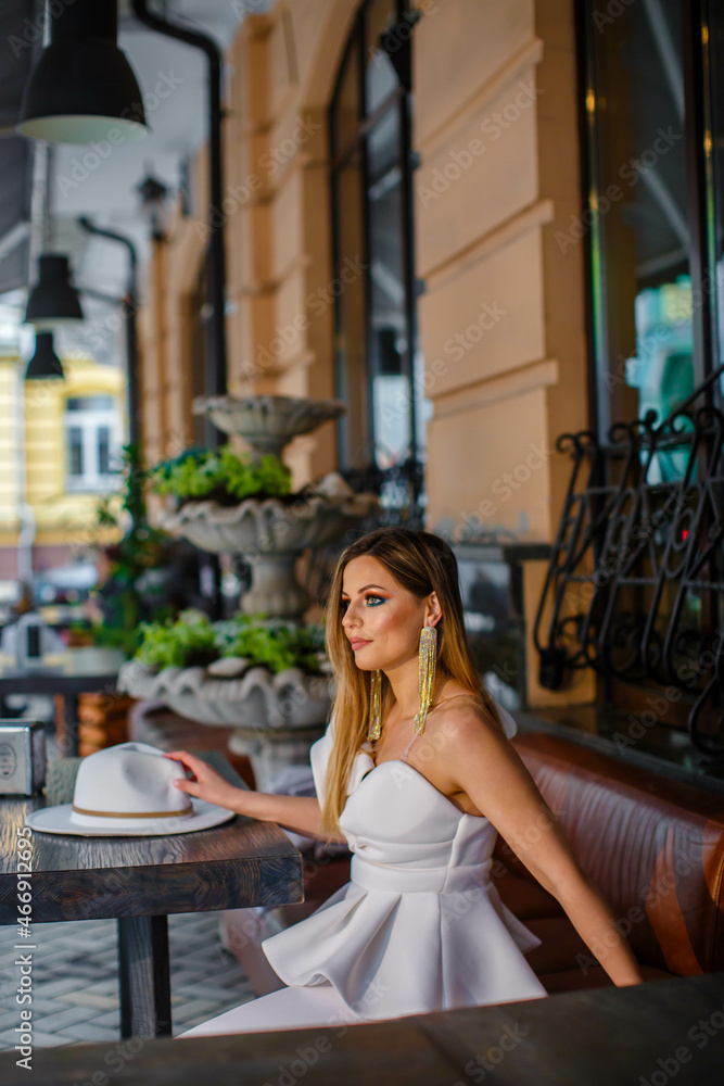 Naklejka premium Close up fashionable woman portrait of young pretty fashionable girl posing in white dress and retro hat while sitting in cafe. Fashion chic