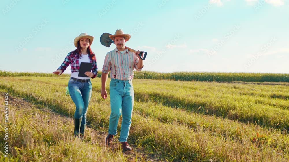 Caucasian woman farmer in a hat walking through the middle of the golden field of wheat with her male colleague and tapping, scrolling and typing on the tablet Caucasian woman farmer in a hat walking through the middle of the golden field of wheat with her male colleague and tapping, scrolling and typing on the tablet