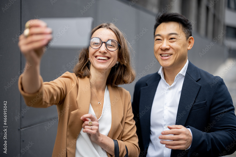 Asian businessman and caucasian businesswoman taking selfie on smartphone on city street. Concept of business cooperation. Idea of modern successful business people. Man waving hand at camera Asian businessman and caucasian businesswoman taking selfie on smartphone on city street. Concept of business cooperation. Idea of modern successful business people. Man waving hand at camera