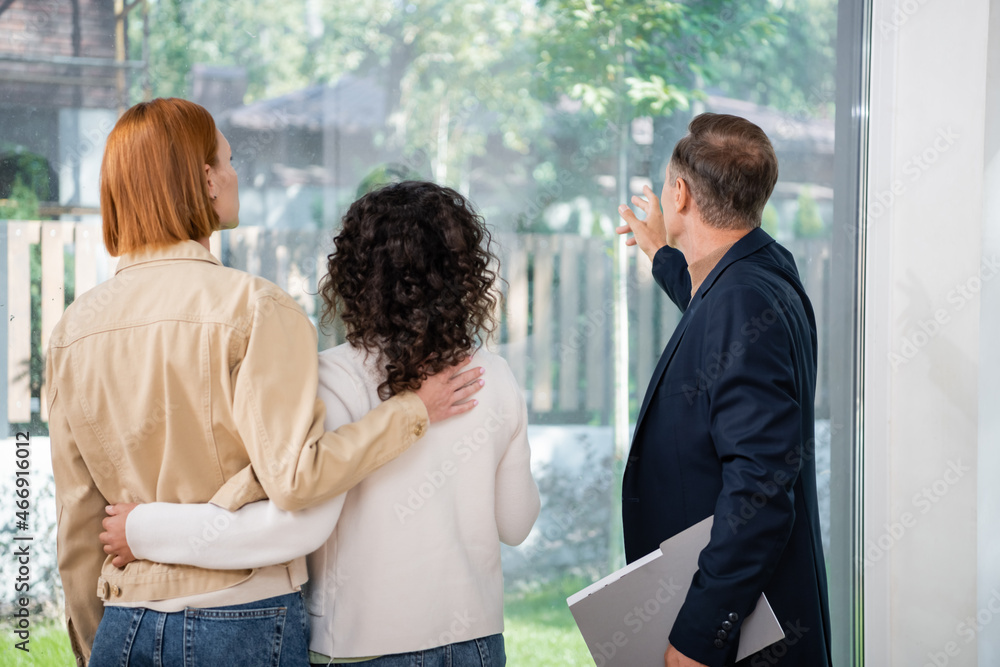 back view of realtor holding folder and pointing at panoramic window near interracial lesbian couple inside of new house back view of realtor holding folder and pointing at panoramic window near interracial lesbian couple inside of new house