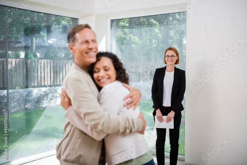 cheerful interracial couple hugging near redhead realtor in glasses holding digital tablet on blurred background