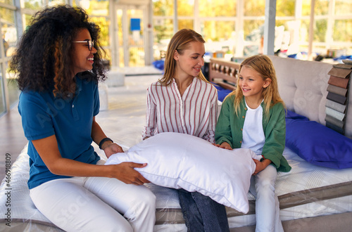 African American saleswoman helps shoppers choose pillow while sitting on bed in store. Mom and her lovely daughter are buying bedding.
