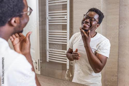 Man Wearing Pyjamas Standing At Sink Putting On Moisturizer In Bathroom. Young man checking his stubble in bathroom. Attractive Well-Shaved African American Man Touching Smooth Face After Shaving photo