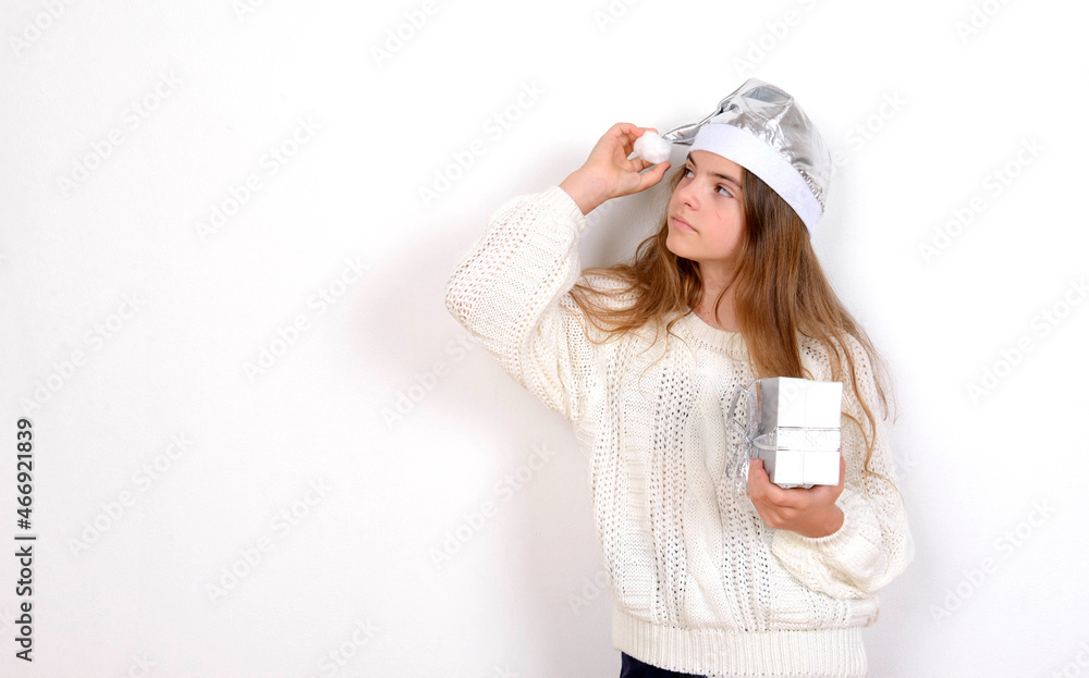 Young girl in white sweater and silver santa hat holds present and touches a popom christmas Young girl in white sweater and silver santa hat holds present and touches a popom christmas