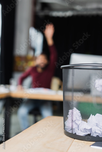 Trash can with crumpled paper on table near blurred african american businessman in office