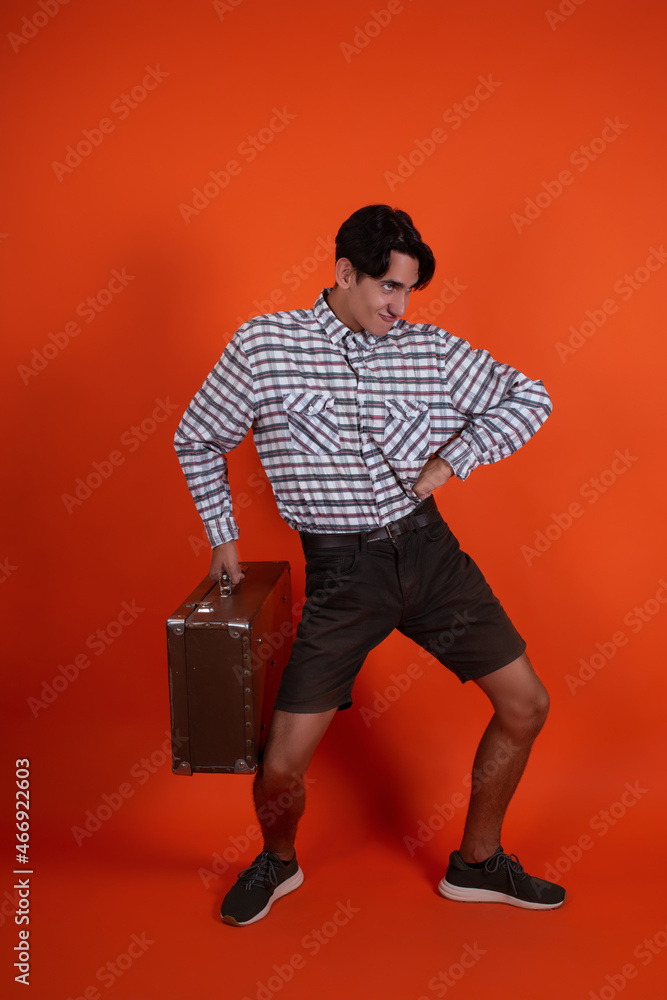 A charismatic handsome guy in a shirt and shorts stands with a suitcase on an orange background. A charismatic handsome guy in a shirt and shorts stands with a suitcase on an orange background.