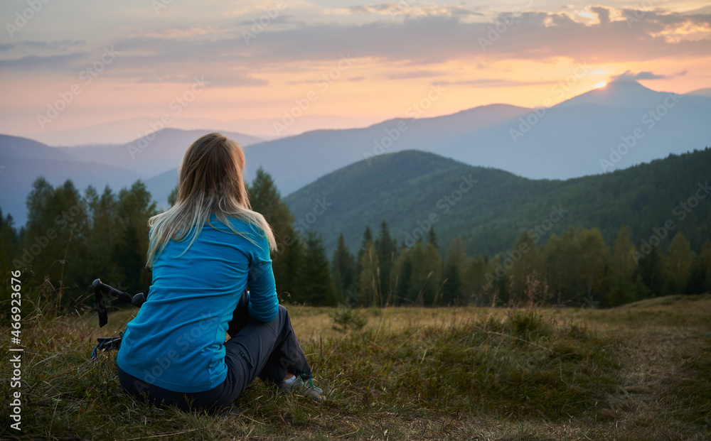 Back view of blonde young woman sitting on grassy hill and enjoying picturesque view of sunrise in mountains. Female hiker resting on grass near trekking poles. Concept of travelling, hiking, nature.