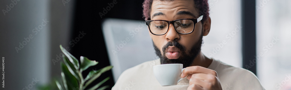 African american businessman in eyeglasses blowing at cup of coffee in office, banner African american businessman in eyeglasses blowing at cup of coffee in office, banner